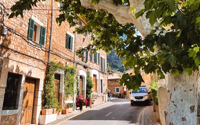 A sunlit village street with stone houses, green shutters, and a leafy tree canopy