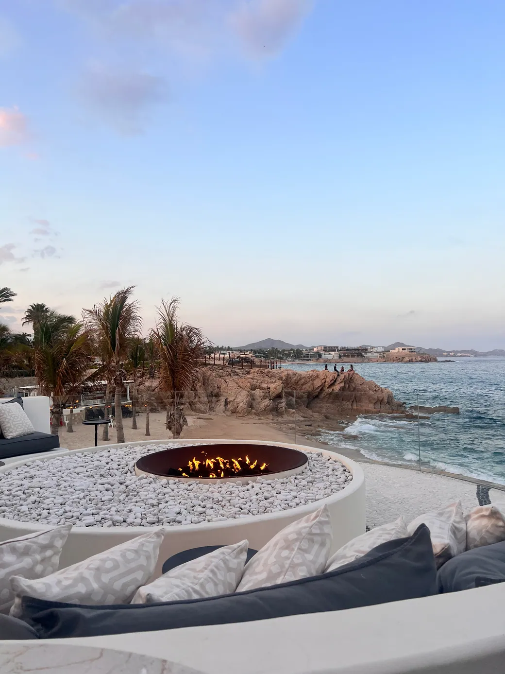 An oceanfront resort terrace at dusk, with a circular fire pit above the water