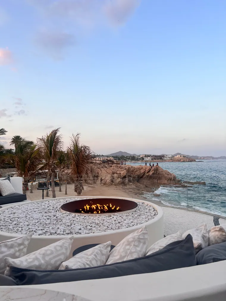 An oceanfront resort terrace at dusk, with a circular fire pit and cushioned lounge seating above the water